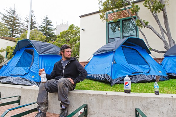 CONFRONTING CITY HALL Aiden, an unhoused resident in SLO for the past five years, sits on a ledge in front of San Luis Obispo City Hall on March 20. Four tents were pitched there as a protest on March 20, just as the city and homeless advocates are working to settle a lawsuit over SLO's treatment of its unhoused population. - PHOTO BY JAYSON MELLOM