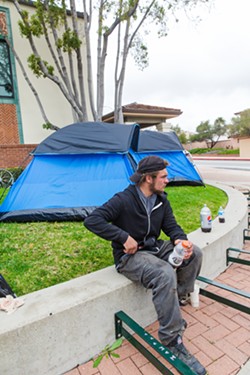 TENT PROTEST Aiden sits beside tents set up on the lawn outside of SLO City Hall on March 20, as the city and homeless advocates work to hash out a settlement in a civil rights lawsuit filed against SLO for the way the city treats its unhoused population. - PHOTO BY JAYSON MELLOM